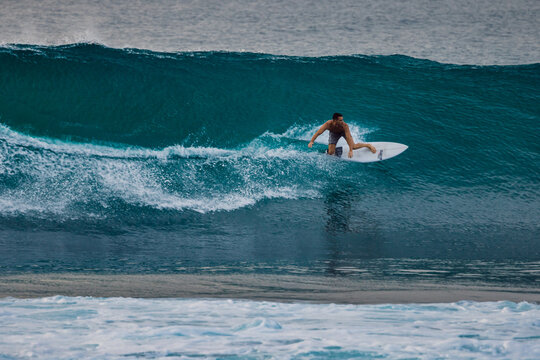 Surfer man crouching low on a white surfboard while carving through a deep blue wave with ocean spray