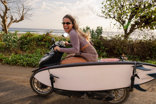 Smiling woman riding scooter with surfboard by the coast
