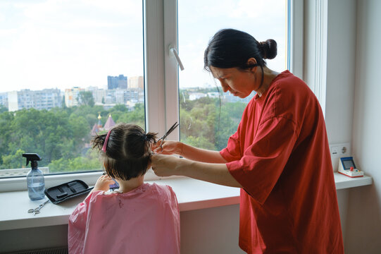 Mother carefully cutting her son's hair by the window.