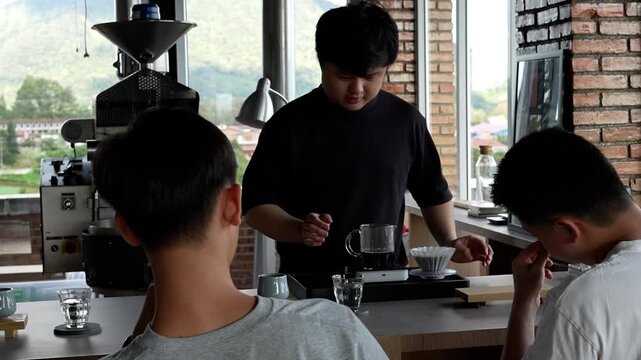 friendly Asian barista smiling at customers while finishing a manual pour-over coffee brew, lifting the dripper at a scenic roastery cafe