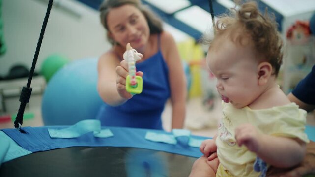 Smiling woman holding a giraffe teether toy for a curly-haired baby girl seated on a round nest swing in a therapy room, French Riviera, France.