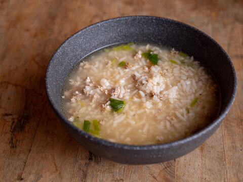 Thai rice soup with minced pork, bok choy and green onion in clear savory broth served in a dark ceramic bowl on a rustic wooden table, comforting breakfast dish