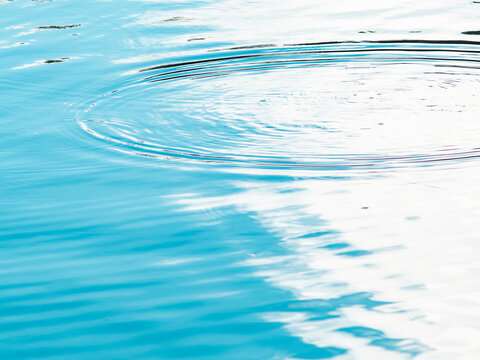Concentric water ripples spreading across calm turquoise blue water surface with white sky reflections, abstract aqua background texture