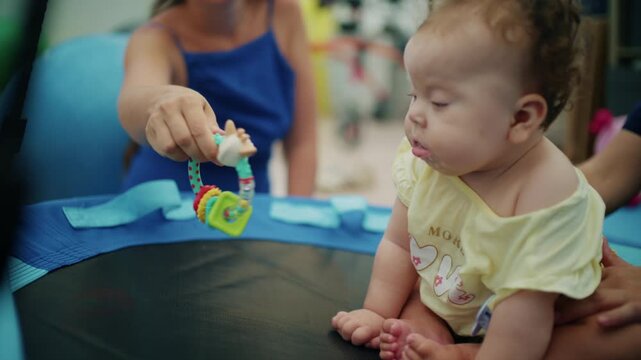 Baby girl on a round therapy swing watching a colorful teether rattle offered by a woman during an early intervention session, French Riviera, France.