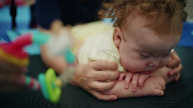 Baby girl resting on her forearms during tummy time on a therapy swing as therapist hands show her a teether toy, French Riviera, France.