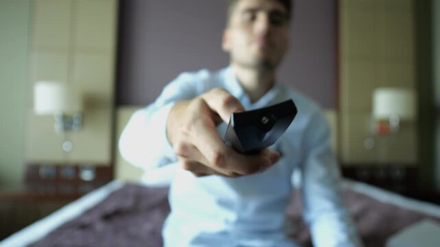 Focus on male hand of young businessman sit on bed switch channels on TV remote control at modern hotel room. Cozy relaxation during business trip showing comfort and leisure with blur background
