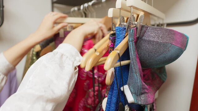 Close-up of businesswoman's hands choosing between colorful sequin dresses, a glittery corset, blouses, and a blue mini skirt in a Spain secondhand shop. Eye-level view of choosing thrift clothes.
