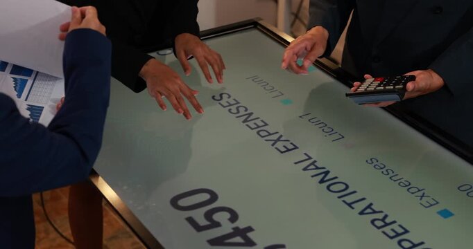 Business people review financial charts and expenses on an interactive touchscreen table while using a calculator during a budget meeting.