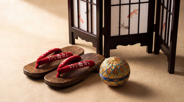 A studio still life featuring a pair of traditional Japanese wooden geta sandals with floral straps, a decorative temari ball, and a miniature shoji screen.