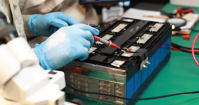 Technician wearing blue gloves tests an electric vehicle battery pack with probes on a workshop bench, showing repair, maintenance and clean energy technology.