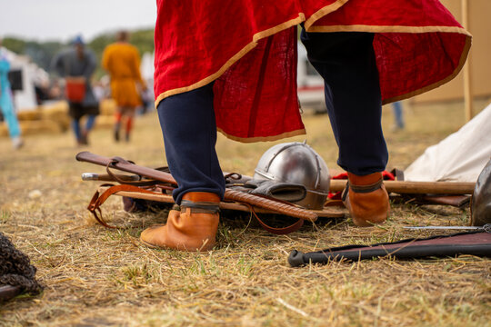 A participant prepares for a medieval reenactment beside a steel helmet and historical gear laid on straw. Costumed figures and tents are visible in the background.