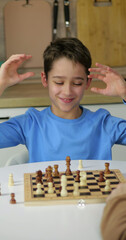 Smiling boy plays chess at home with hands raised above board. Young strategist enjoys thoughtful game during leisure time. Indoor scene shows focus, joy, and learning through competition. © julia_diak