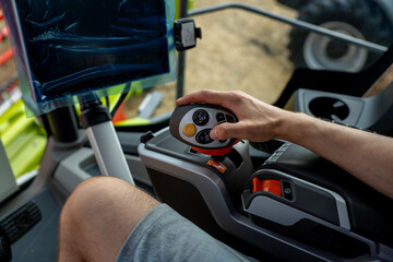 An operator uses a joystick inside an agricultural machine cab, with control buttons and a monitor visible beside the seat. © Marina