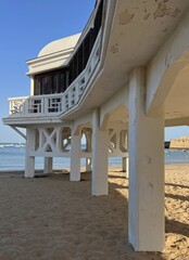 Architectural view beneath the historic Balneario de la Palma building on La Caleta beach in Cadiz, Spain, showing its elevated structure, supporting pillars and sandy shoreline. © MaruStock