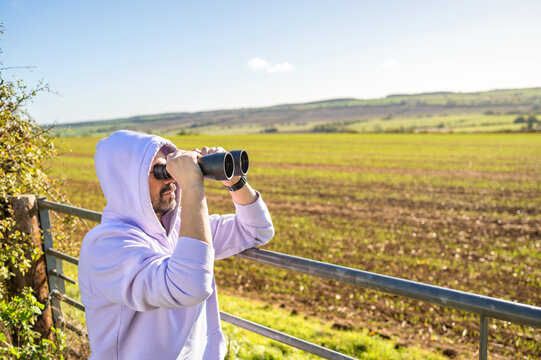 Man in hoodie looking through binoculars over rolling countryside from a rural observation viewpoint. bird watching