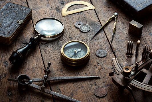 Antique navigation and exploration still life centered around an old compass on a rustic wooden surface, surrounded by an astrolabe, dividers, antique book, coins, spyglass, and magnifying glass. 