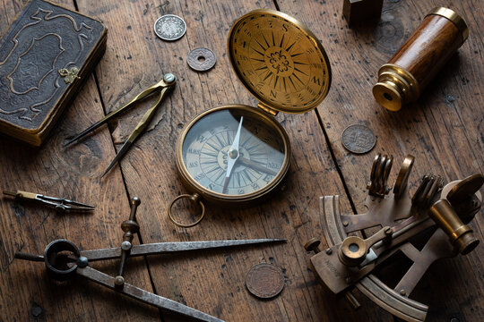 Antique navigation and exploration still life centered around an old compass on a rustic wooden surface, surrounded by an astrolabe, dividers, antique book, coins, spyglass, and magnifying glass. 