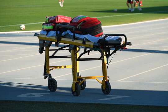 Emergency stretcher with medical bags beside soccer field. Sports venue first aid and ambulance response equipment. Athletic injury care and emergency preparedness. Football training ground