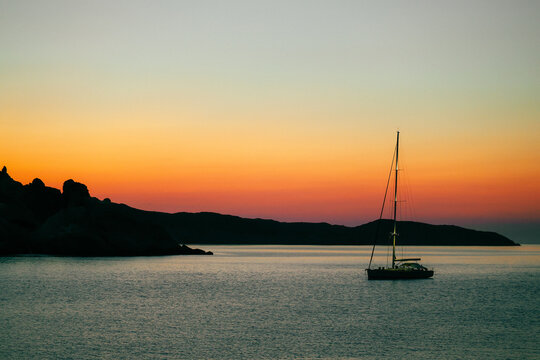 The silhouette of a sailboat during a beautiful sunset at Mandrakia on the island of Milos in Greece. The Greek sky is orange and red as the sun descends behind the rocky coastline. 