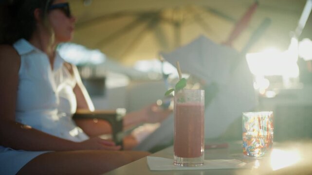 Pink cocktail with mint and a colorful glass tumbler on a table as a mother sits beside a baby stroller in golden backlight, French Riviera, France.