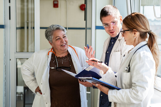 Three doctors are getting ready in the hospital hallway before heading to a meeting. They talk briefly and review their notes as they prepare to discuss their work together.