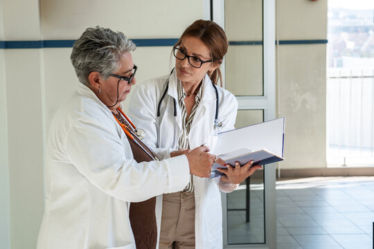 Two female doctors, one senior and one younger, discuss a case in the hallway outside a medical office. They exchange ideas and review information together in a calm and professional setting.