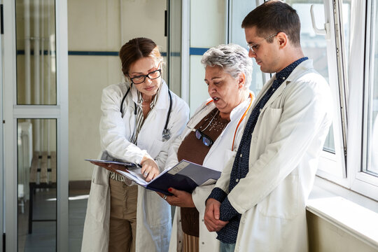 Three doctors are getting ready in the hospital hallway before heading to a meeting. They talk briefly and review their notes as they prepare to discuss their work together.