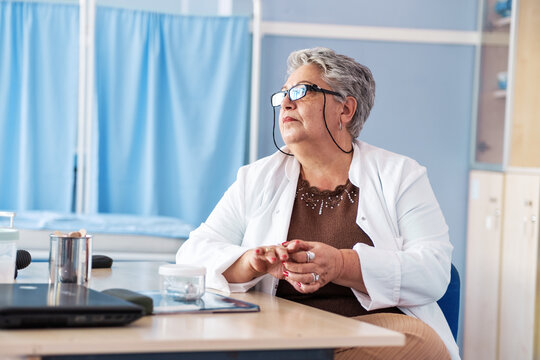 An older female doctor stands in her medical office, looking professional and confident. The portrait reflects her experience and dedication to caring for patients