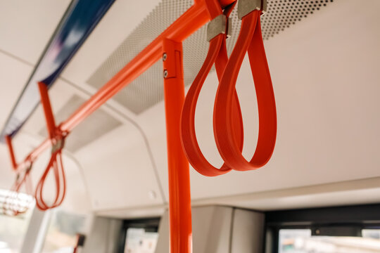 Close up of red hanging hand straps on an orange handrail inside a modern tram, public transport interior, empty vehicle, daylight from windows, city commute, grainy film texture, high quality photo