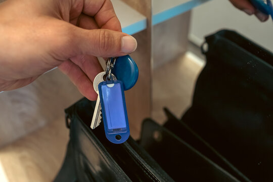 Close up of a hand holding a set of keys with a blue key tag and round fob above an open black leather bag on a wooden shelf, putting keys away, everyday routine, grainy film texture, high quality pho