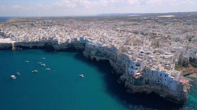 Polignano a Mare Puglia Italy aerial view of white town on cliff and beach. Drone approaching Lama Monachile cove, steep limestone rocks, moving towards yachts and moored boats in summer