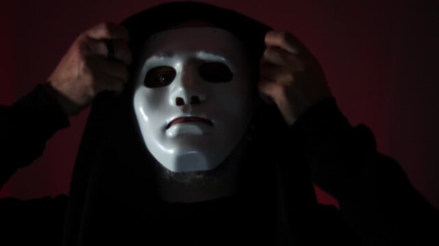 Atmospheric close up of a hooded person with a white mask for halloween, white under dramatic red backlighting