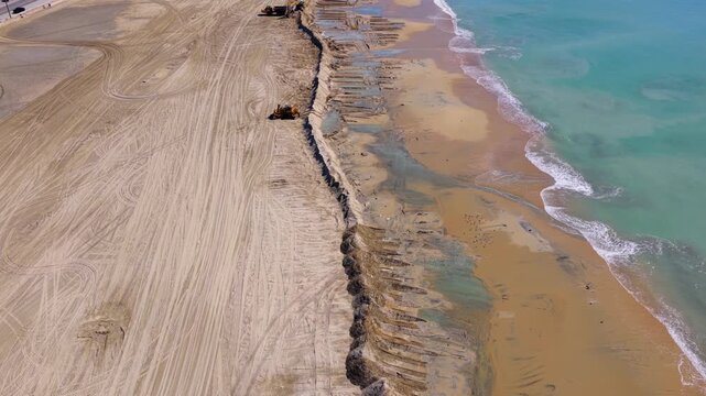 Long Beach, California, USA - October 15, 2026: Bulldozer, excavator and dump truck build a long winding sand berm between the beach and the surf, gulls on the wet sand, after Tropical Storm Rachel.