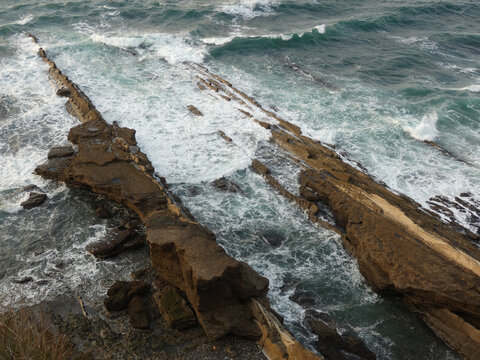 High-angle view of jagged coastal rock ridges surrounded by swirling white surf and textured blue-green ocean water
