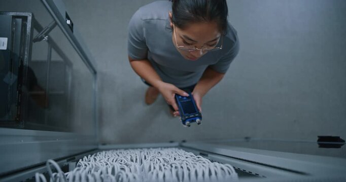 Female Network Engineer Verifies Ethernet Connection Using Portable Cable Analyzer Connected to Patch Cord. Open Server Rack Reveals Dense Cabling, Highlighting Physical Layer Testing and Diagnostics.