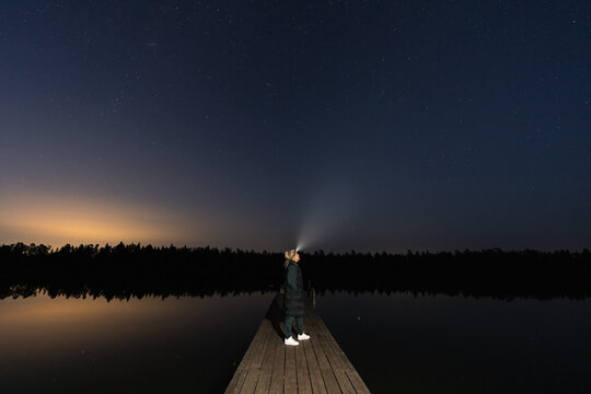 Woman with headlamp standing on wooden dock over calm lake looking at starry sky and distant light pollution during blue hour night.