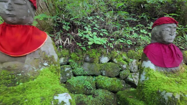 Stone Jizo statue wearing a red bib and cap sitting on mossy stone in the Kanmangafuchi Abyss forest, Nikko, Tochigi Prefecture, Japan.