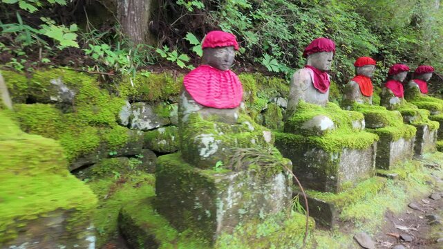 A row of moss-covered stone Jizo statues with red hats and bibs along a path in the Kanman-ga-fuchi Abyss in Nikko, Tochigi, Japan.