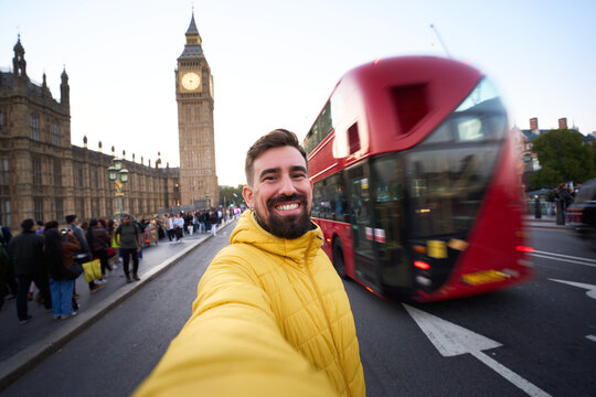 Smiling caucasian man taking a selfie in London with Big Ben and a red double decker bus passing behind. Wide angle portrait on Westminster Bridge captures city energy, travel joy and urban vibes.