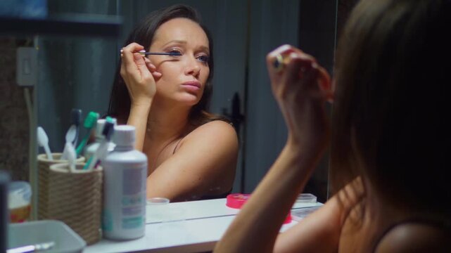 Woman applies mascara in dim bathroom light. She looks tired, focused on her eyes. Mirror reflects her careful, deliberate motion. Soft lighting enhances intimate, personal moment
