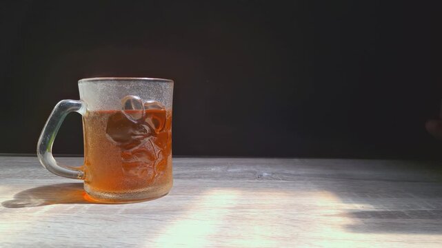 Golden liquid fills textured glass mug. Hot amber tea with steam rising on a wooden table in warm morning sunlight against a dark background. Ideal for a calm breakfast and a moment.