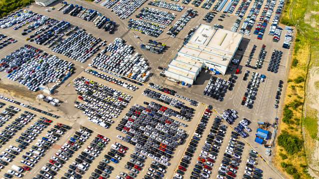 High angle perspective of cars in distribution terminal. Rows of various colored automobiles parked systematically in a large open-air lot adjacent to a warehouse facility.