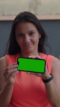 Softly smiling woman holding smartphone with green screen, outdoor app display