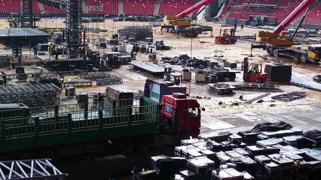 Cranes and construction crews assemble concert infrastructure inside a stadium lined with empty red seats.