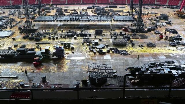 Cranes and construction crews assemble concert infrastructure inside a stadium lined with empty red seats.