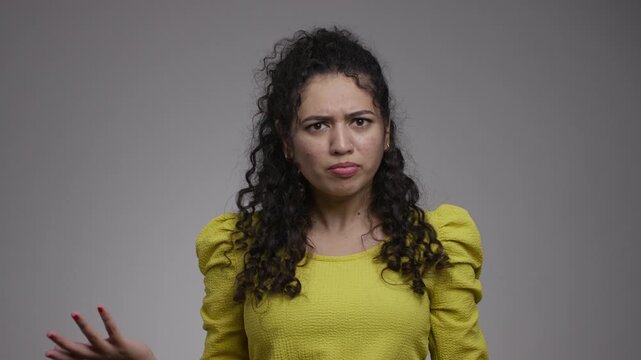 A South Asian woman raises both hands toward herself, furrows her brows and gestures with one hand before looking down.