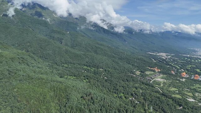 Aerial drone footage of green forested mountains with white clouds above a valley town, Southwest China, D-Log lossless file format