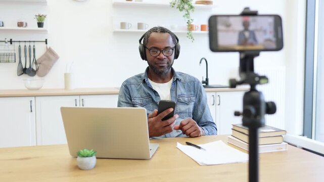 An African American creator smiles and gestures while recording at home, using a smartphone setup beside a laptop, notes, and books