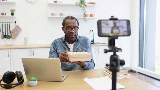 A creator presents a small package at a kitchen table while a phone records, with a laptop, shipping materials, and home shelves nearby