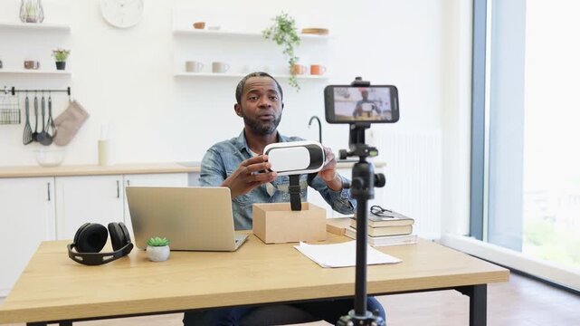 An African American creator presents a small white device at a desk, while a phone records him beside a laptop, headphones, papers, and a shipping box